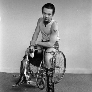 Photograph: a youngYoung man in a wheelchair with the left foot on the floor. He stares into the camera with a direct gaze and holds a coors beer in his right hand. He wears a wife beater t shirt and cut-off levis so that we see his leg braces and tattoo of a bird on his left arm.