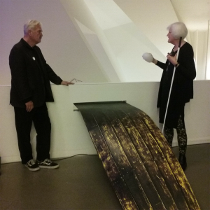 The other side of The Enchanted sculpture features a long strip of wooden planking that resembles a boat protruding out from a wall. A man stands to the left and a woman with white hair stands to the right with a cane, holding a white, rubbery object in her right hand