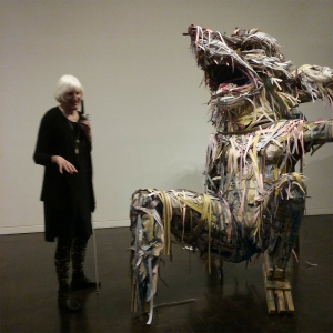 On the left a tall white-haired woman stands dressed in black and holding a white cane, to her right a sculpture made of strips of paper and cardboard of a fox like creature, leaning back on its hind legs and one front leg resting behind its body, appearing to laugh, and towering over the woman.