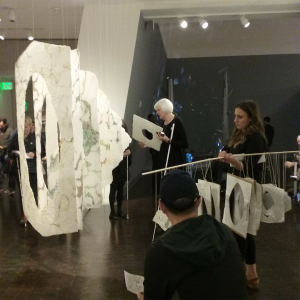 Sheets of exquisite and handmade paper with complex textures, pale colors and central holes hang from the ceiling in a row. A tall woman with white hair and white cane holds a piece of paper. A woman to her right holds a pole with a smaller version of the sculpture. In the background a woman reads from a Braille folder and several people holding canes listen to the conversations.