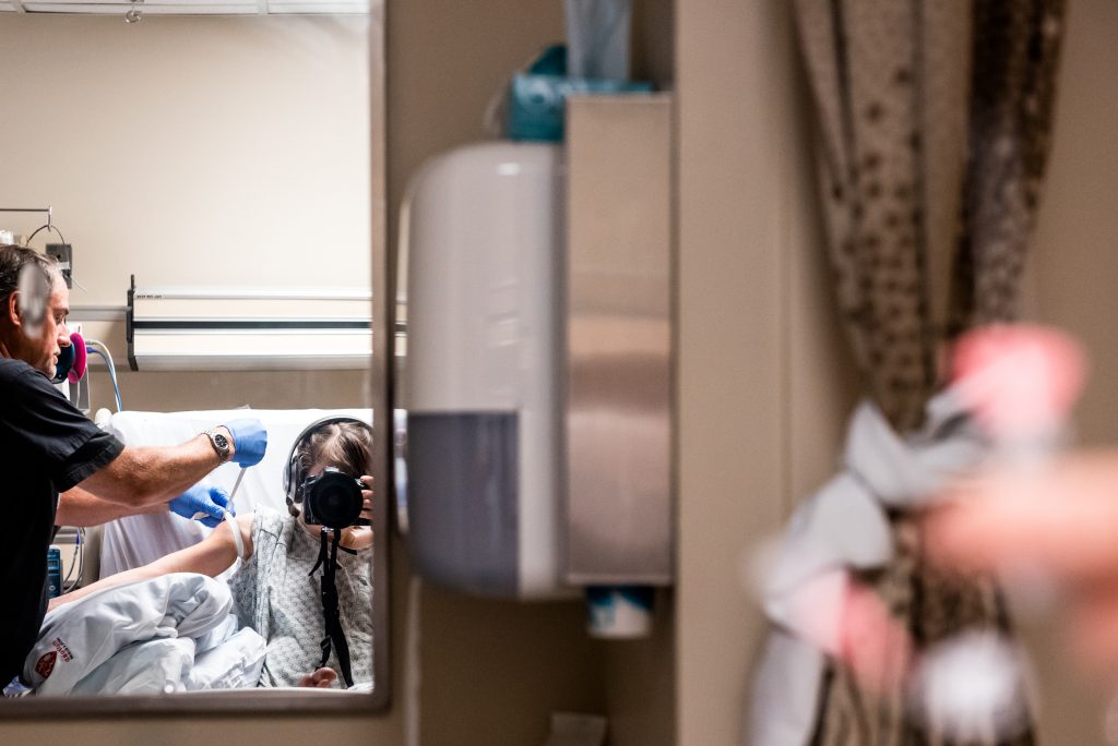 White woman (author) holds black camera up to face in hospital gown.