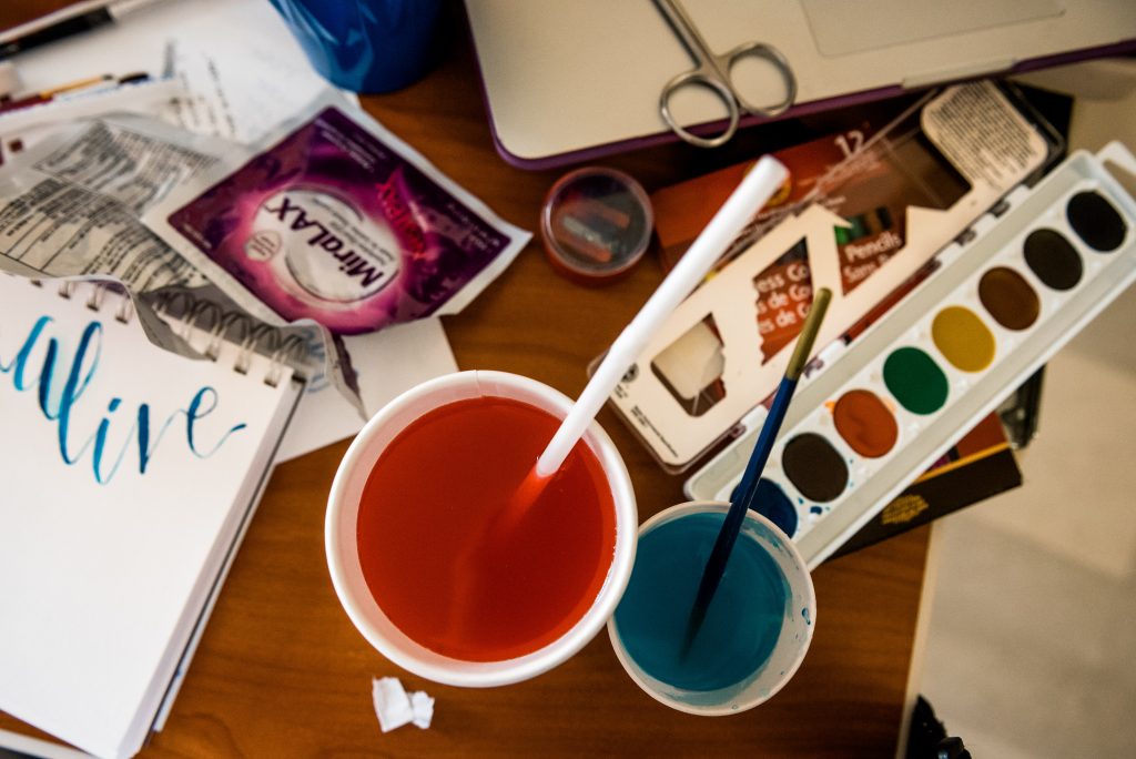 Water color paints, colored water cups with brushes and a sketchpad sit on a table amongst medical wrapper and papers.