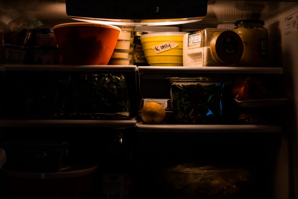 Dark inside of refrigerator shows outlines of containers and light shows yellow container with labeled tape.
